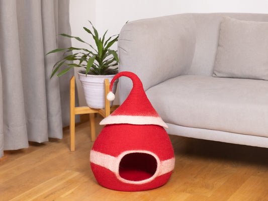 Red pet bed shaped like a Santa hat on a wooden floor with a couch and plant in the background.