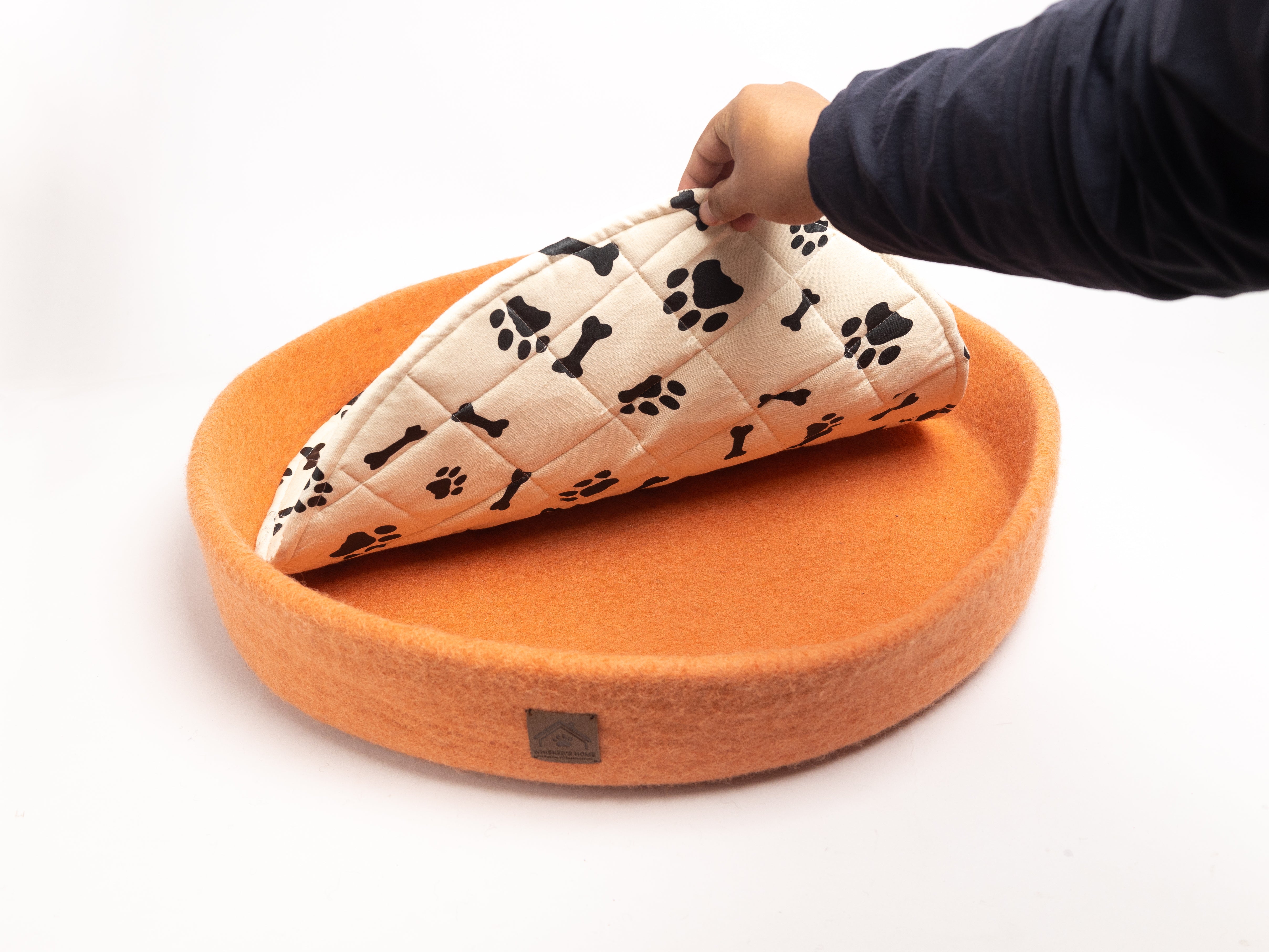 orange colored round wool dog bed with black paw and bone printed in white cushion, person's hand lifting the cushion