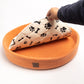 orange colored round wool dog bed with black paw and bone printed in white cushion, person's hand lifting the cushion
