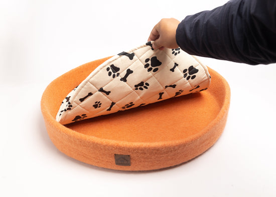 orange colored round wool dog bed with black paw and bone printed in white cushion, person's hand lifting the cushion