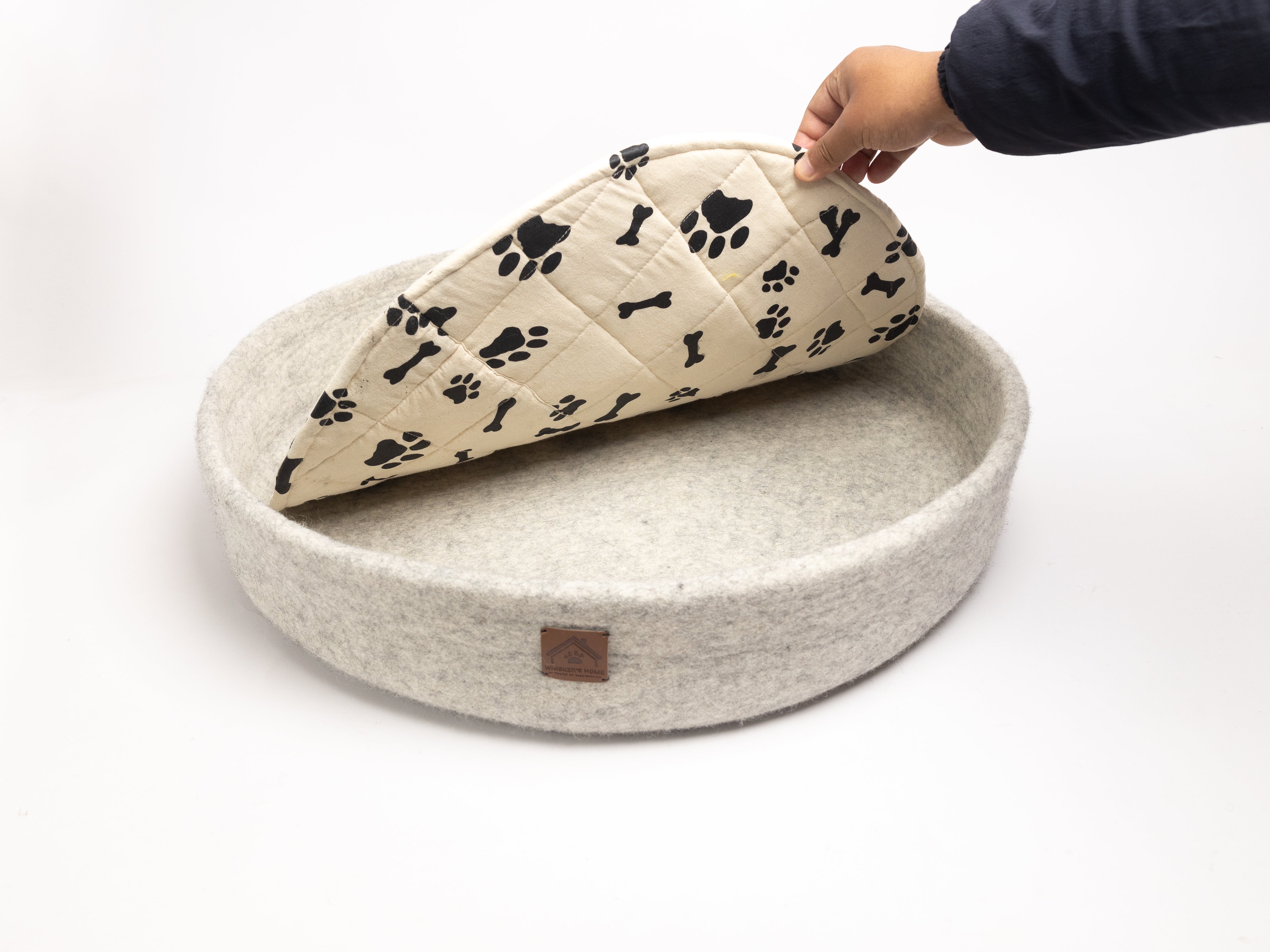 grey felted dog bed with black paw print patterns, round shape, person's hand holding cushion