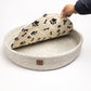 grey felted dog bed with black paw print patterns, round shape, person's hand holding cushion