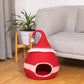 Red pet bed shaped like a Santa hat on a wooden floor with a couch and plant in the background.