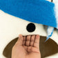Close-up of a plush toy with a blue hat and brown mouth, held by a hand against a white background.